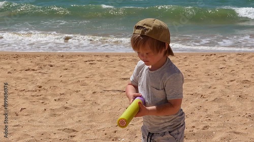 Cute Little Boy in Cap and Grey T-Shirt Playing with Water Gun on Ocean Beach