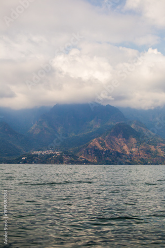 Calming lake reflects distant, mist-covered mountains. Clouds drift above rugged, green-hued peaks. Water ripples gently under soft, overcast skies. Lake Atitlan, Guatemala.