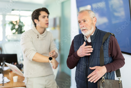 Quadro su tela Young man sternly scolds elderly male colleague for being late for work in offic