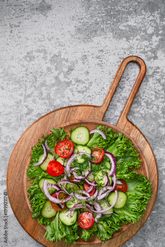 Fresh summer salad with cherry tomatoes, cucumbers and red onion on a bed of green lettuce leaves, served on a wooden cutting board, top view with copy space.