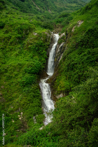 Tall cascading waterfall flowing down a steep lush green mountain ravine
