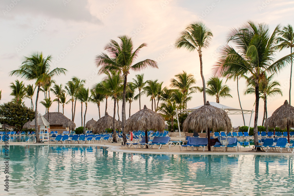Fototapeta premium Tropical resort pool area with palm trees and sun loungers at sunset