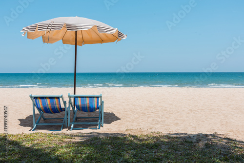 Two colorful beach chairs under striped umbrella on sunny tropical beach