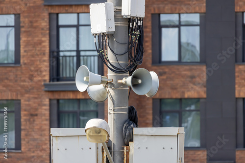 Public address system with multiple loudspeakers on metal pole against brick building. Outdoor siren and alert equipment for emergency notifications in city.
