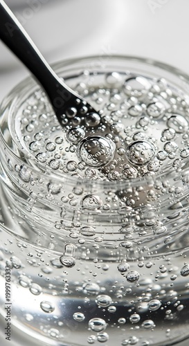 Clear viscous substance in a container displays numerous trapped air bubbles being disturbed by a small dark utensil.