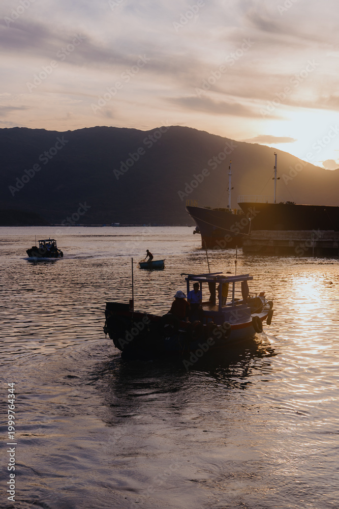Obraz premium Fishermen on a boat head out to sea at sunrise in Quy Nhon, Binh Dinh, Vietnam. The sun rises behind the mountains, casting a golden glow on the water.