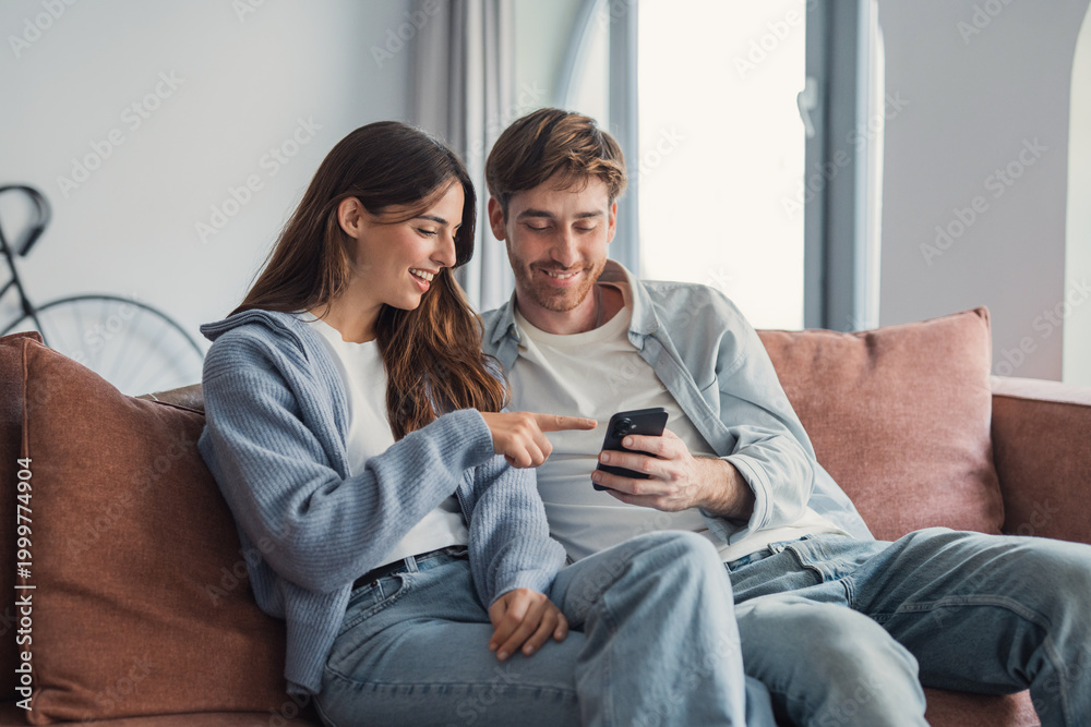 Obraz premium Young couple sitting on a sofa at home smiling and sharing a smartphone while relaxed together
