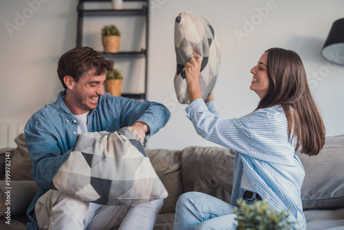 Fotografi A young couple share a playful pillow fight on their living room couch, laughing and enjoying a cozy, relaxed moment together that captures intimacy, fun and everyday relationship joy