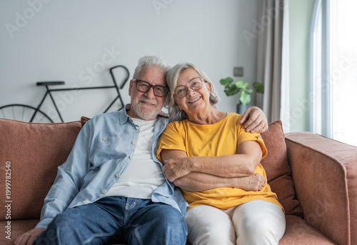 Elderly couple sits close on a comfortable sofa in their living room, smiling and embracing with warmth and affection. Relaxed, happy retirees enjoying companionship, comfort, and quiet domestic life.