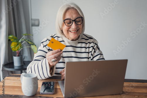Konstfotografi Smiling senior woman sits at a wooden table in her living room using a laptop and credit card to make an online purchase, enjoying technology, convenience and independent everyday life