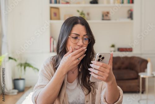 Foto A surprised woman wearing glasses reads a message on her smartphone while sitting in a cozy living room, reacting emotionally to unexpected news or a notification during her time at home