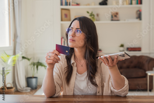 Photos Frustrated young woman at home holding a credit card and smartphone, hesitating during an online payment