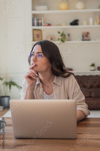 Slika na platnu A pensive young woman with glasses sits at a wooden desk using a laptop in a cozy living room