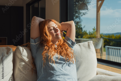 Fotografia Red-haired woman lounges on a sofa with hands behind her head, basking in warm sunlight through large windows and enjoying a calm, peaceful moment of relaxation at home