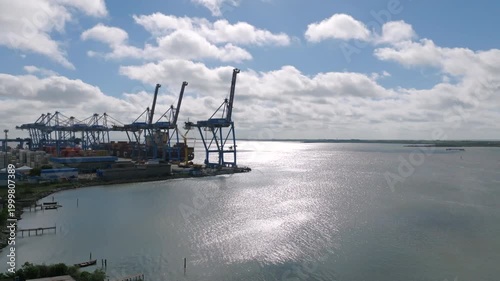 Aerial pan revealing container port terminal with towering cranes and stacked cargo containers from open estuarine waterway
