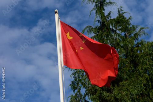 A vibrant red flag of China waving against a backdrop of blue skies and green trees
