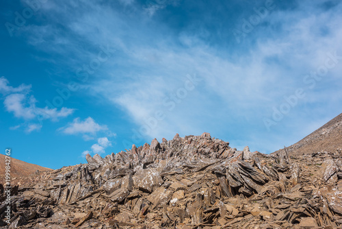 Sunlit stone hill in high mountain valley under clouds in blue sky. Pile of s...