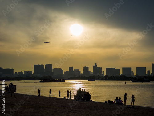 A scene featuring the coastal skyline with the silhouettes of people relaxing on the city beach at golden hour (Minato, Tokyo, Japan)