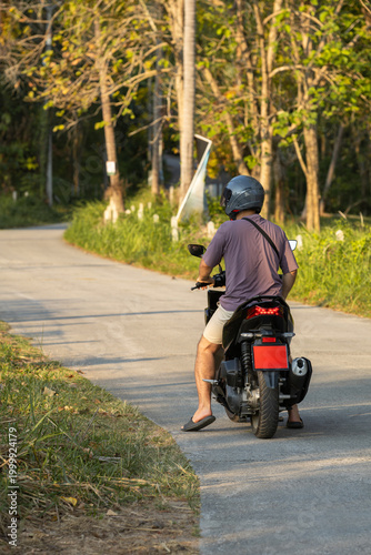 Rear view of a man wearing a helmet and sandals, riding a black scooter on a winding sunny rural road in Asia, with lush greenery and trees creating a warm, relaxed travel atmosphere.