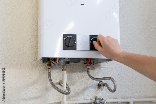 Person's hand turning a knob on a white wall-mounted gas water heater, demonstrating installation, maintenance, or repair work on heating equipment and home appliances