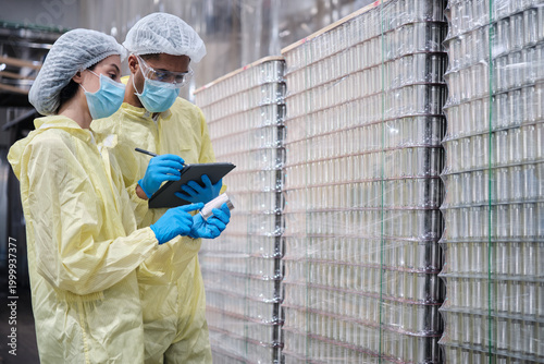 Two industrial engineer supervisors inspect and examine quality of stainless steel containers, production process in canned food manufacturing factory, and protective uniforms for hygiene operations.