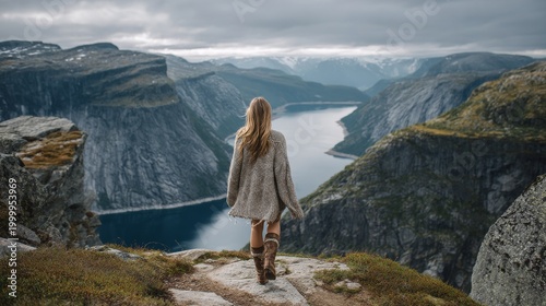 Rear view of a woman in warm sweater and boots standing on a rocky mountain viewpoint above a deep fjord, surrounded by steep cliffs and cloudy sky