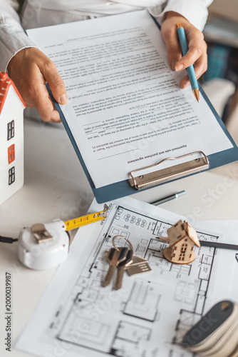 Professional female realtor explains housing terms client while preparing contract in bright office. Real estate transaction with apartment plans, model house and trusted mortgage assistance concept.