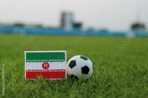 Slika na platnu Embroidered Iran national flag next to a classic soccer ball on green grass pitch