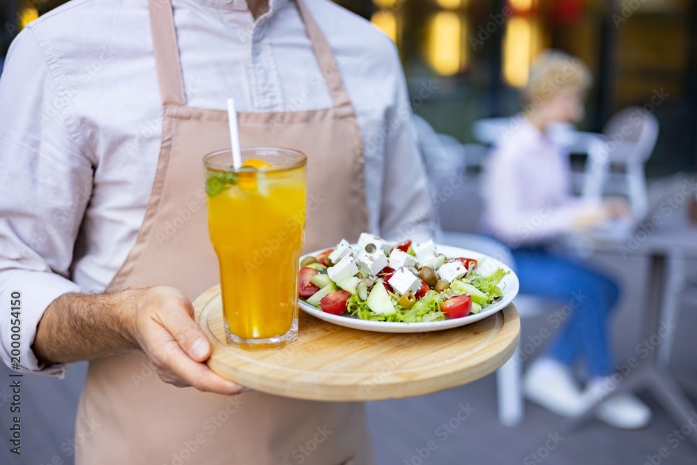 Fototapeta premium A server in an apron presents a fresh Greek salad and a refreshing orange beverage on a wooden tray at an outdoor cafe