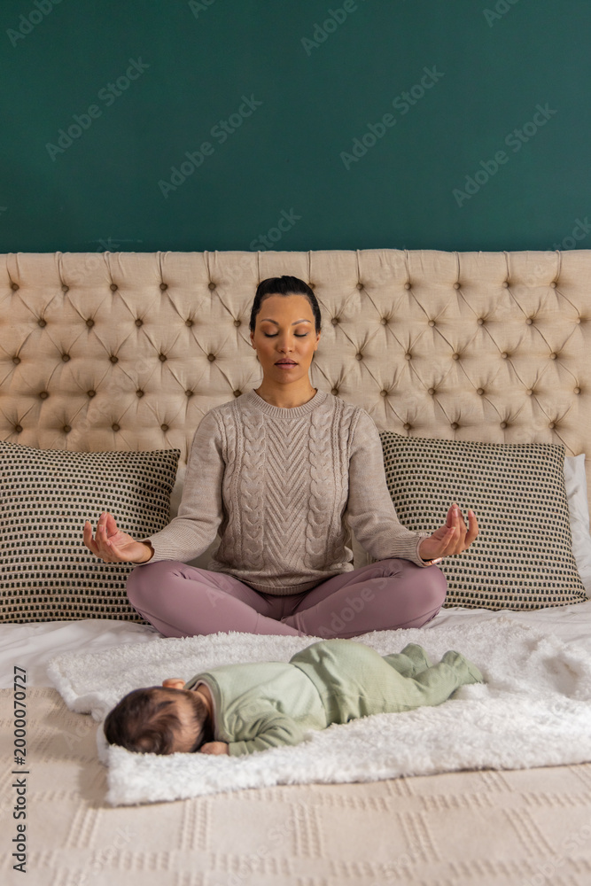 Obraz premium Mid-adult mother and infant daughter sitting meditating on bed with headboard, knit sweater, onesie