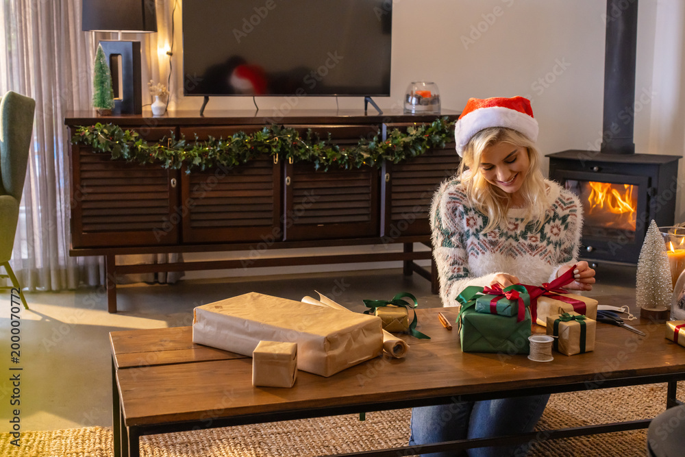 Fototapeta premium Woman wearing Santa hat and patterned knit sweater wrapping kraft paper gifts in living room
