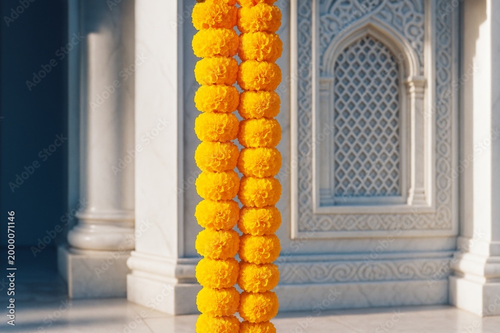 Fototapeta premium Bright yellow marigold flower garland hanging in sunlight against ornate stone architectural background with intricate patterns and columns. Ai generative