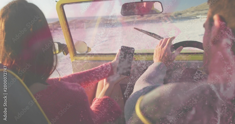 Fototapeta premium Driving couple wearing pink sweater and gray jacket on sand in yellow buggy, holding smartphone