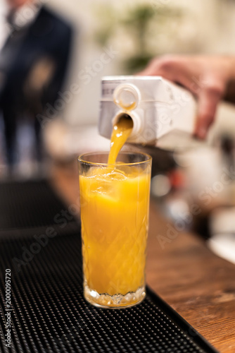 Close-up of orange juice being poured from a carton into a tall ice-filled glass on a bar mat during event drink service