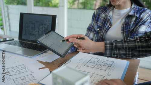 Architects reviewing house floor plans at desk, laptop and tablet in bright office, collaborating on home design project with blueprints and documents.