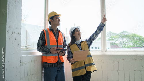 Two construction workers in safety vests and hard hats inspect an interior near a large window, one pointing while holding a clipboard, suggesting building site planning and teamwork.