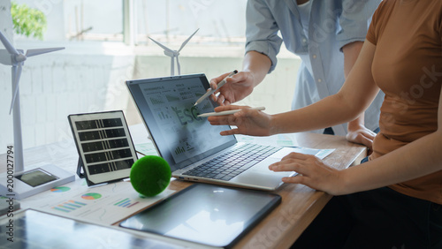 Two colleagues discuss ESG data on a laptop in a bright office with wind turbine models, tablet and reports, suggesting sustainable business, renewable energy planning and green finance.