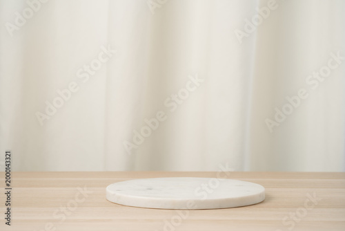 Minimal white marble pedestal placed on a light wooden table with a soft neutral curtain backdrop, ideal for product display, studio mockups, presentations, and clean commercial staging.
