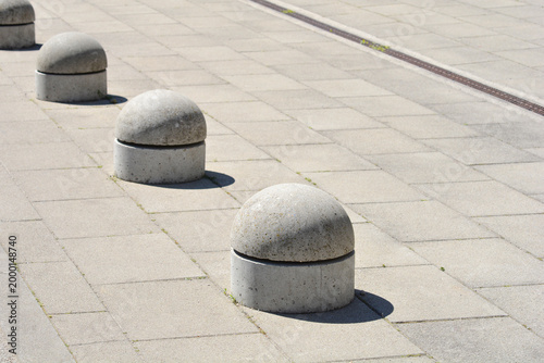 A line of grey spherical concrete bollards stands on a paved city sidewalk in bright sunlight casting soft shadows