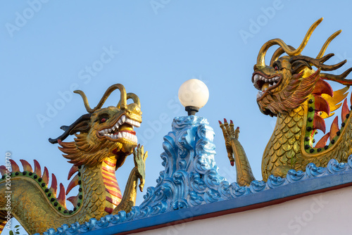 Two magnificent golden dragon statues with intricate details adorn the roof of a temple, set against a clear blue sky. These powerful symbols are often seen in Asian religious architecture.