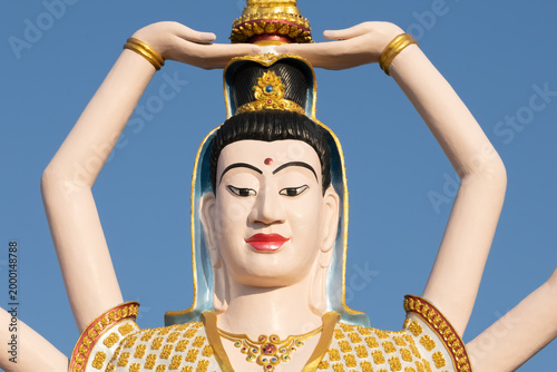 Close-up of a multi-armed Buddhist deity statue with an ornate golden headdress and serene expression against a bright blue sky, highlighting intricate details and spiritual tranquility.