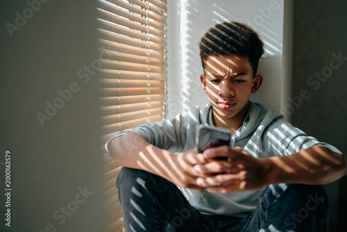 Teenager texting on smartphone near window with sunlight casting shadows, thoughtful expression and calm atmosphere, authentic lifestyle