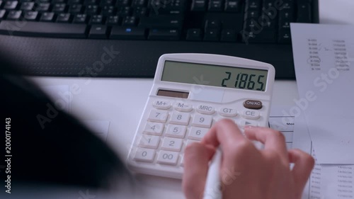 Focused person calculating business finance paperwork using white calculator near computer keyboard examining tax document and accounting report on modern office desk showing concentration