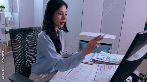 Young focused asian businesswoman sitting at modern office desk pointing computer monitor analyzing document writing paperwork working late night corporate task with serious professional dedication