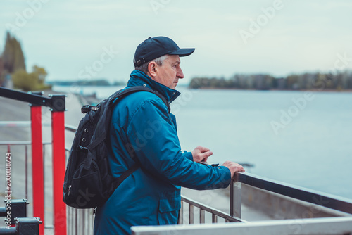 Thoughtful Mature Man with Backpack Looking at River. Active Lifestyle and Travel Concept
