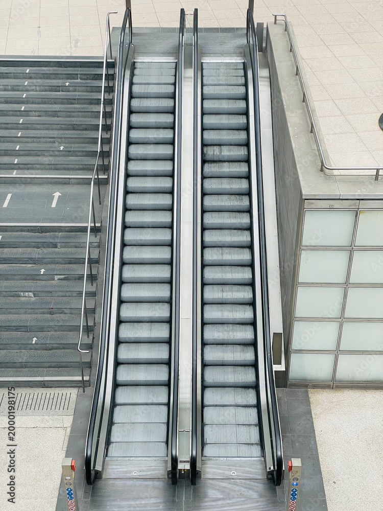 Obraz premium Double modern escalators and stone stairs in urban public transport hub. High-angle shot showing two parallel escalators next to a wide stone staircase in a contemporary public space
