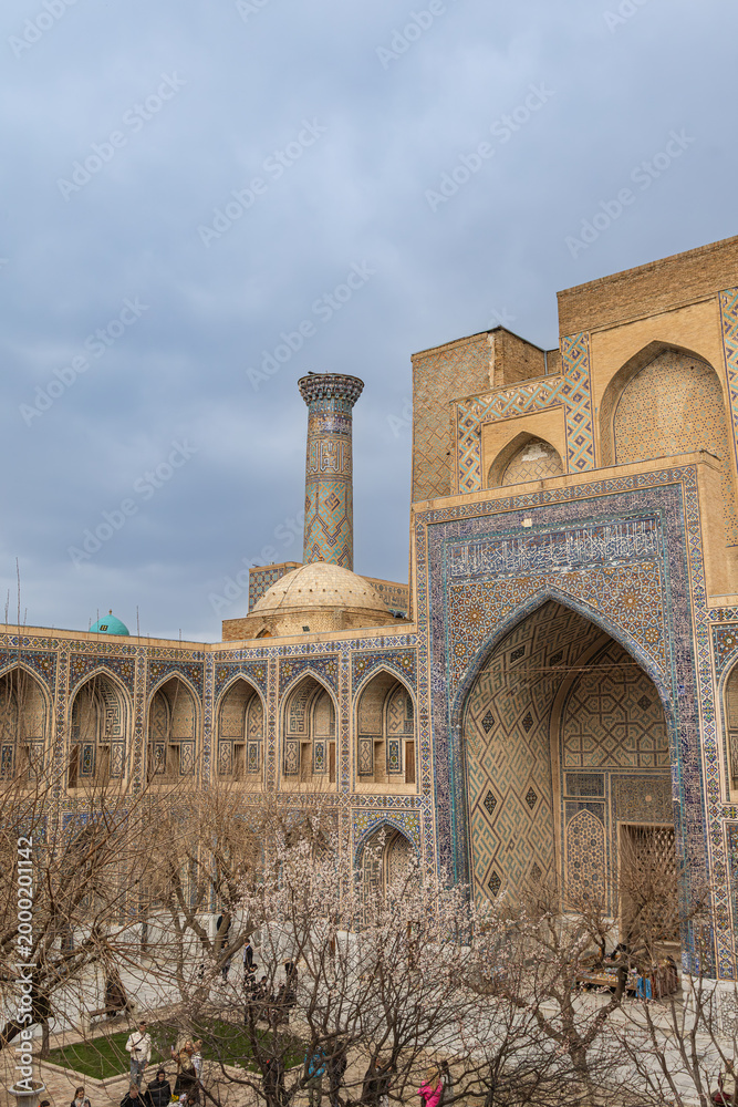 Behang View of Ulugbek Madrasa courtyard with Islamic tilework, pointed archways, a tall decorated minaret and blossoming trees; a historic travel landmark showcasing  architectural heritage of Registan