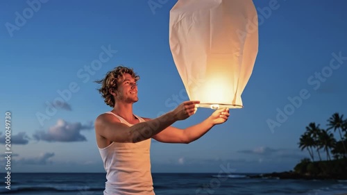 Young man launching a sky lantern on a tropical beach at twilight with palm trees and ocean waves in the background