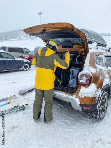 Skier preparing gear beside snowy car in busy winter mountain parking lot.
