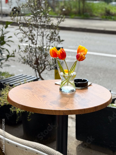 Round wooden cafe table with tulips in glass vase placed on outdoor terrace. Minimal dining setup, street cafe atmosphere, relaxation and everyday urban lifestyle.